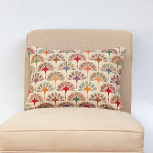 May include: Rectangular cushion featuring a repeating fan design in red, orange, green, blue, and purple on a cream backdrop. The cushion is placed on a beige upholstered chair.
