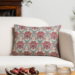 Op de afbeelding: Rechthoekig decoratief kussen met een bloemmotief. Het kussen heeft een herhalend ontwerp van gestileerde bloemen in rood, blauw en beige. Het ligt op een witte bank, wat wijst op een interieurthema.