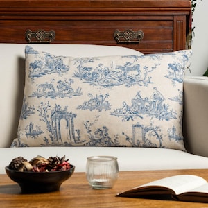 May include: A rectangular throw pillow featuring a blue and white toile de Jouy pattern on a beige fabric background. The print depicts various pastoral scenes including figures, animals, trees, and architectural structures across the surface.