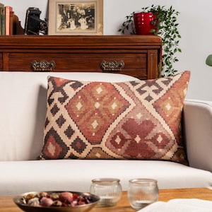 May include: Rectangular cushion with a geometric pattern in shades of red, brown, and beige. The cushion is placed on a white sofa. The design features diamond and square shapes.
