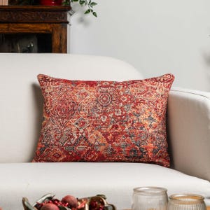 Peut inclure: Coussin rectangulaire décoratif avec un motif détaillé de style vintage dans des tons de rouge, orange et bleu. Le coussin est posé sur un canapé blanc, mettant en valeur son design complexe et sa palette de couleurs. Le coussin a un style de tapis oriental traditionnel.