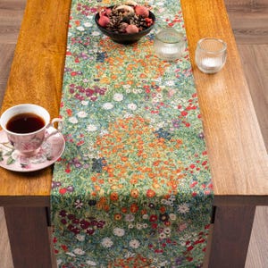 May include: A rectangular table runner featuring a dense, colorful floral pattern in shades of green, orange, red, and white. A white ceramic teacup with a floral design, a black bowl filled with pinecones, and two clear glass votive holders.