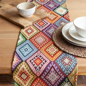 Può includere: Un runner da tavolo colorato con un motivo geometrico in tonalità di rosso, blu, verde, giallo e arancione. Il runner è realizzato in tessuto tessuto e ha un bordo con frange.