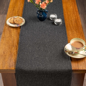 Puede incluir: Un camino de mesa gris oscuro con diseño de espiga sobre una mesa de madera. La mesa está puesta con una taza y platillo florales, un plato de galletas, un jarrón azul con flores y dos candelabros pequeños.