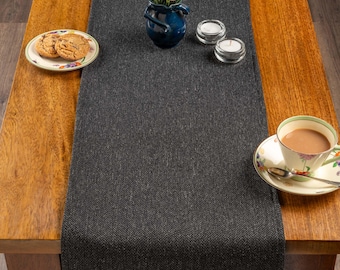 May include: A dark gray herringbone table runner on a wooden table. The table is set with a floral teacup and saucer, a plate of cookies, a blue vase with flowers, and two small candle holders.