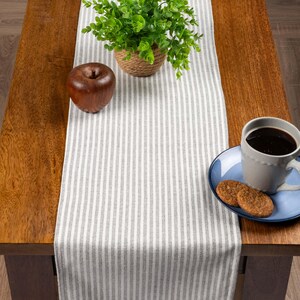Può includere: Una tovaglia a righe grigie e bianche su un tavolo di legno. Una piccola pianta in vaso, una mela di legno, un piatto blu con biscotti e una tazza di caffè sono disposti sul tavolo. La tovaglia aggiunge un tocco decorativo.
