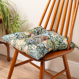 Puede incluir: Un cojín de asiento cuadrado con un patrón de tapiz tejido de follaje verde oscuro, flores color crema y pequeñas bayas rojas. El cojín está acolchado con costuras visibles e incluye lazos de tela en las esquinas para sujetarlo a una silla.