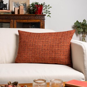 Pode incluir: Uma almofada retangular laranja com um padrão de tecido texturizado repousa sobre um sofá branco. A cor da almofada é um tom quente e terroso, adicionando um toque de cor aos móveis de tons neutros. A almofada é um acessório decorativo para casa.