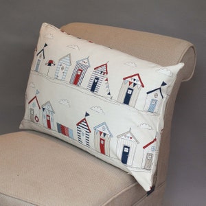 Peut inclure: Un oreiller blanc avec un motif de maisons de plage colorées aux toits rouges et aux portes bleues. Les maisons sont disposées en rangée et sont décorées de drapeaux et d'autres éléments. L'oreiller est sur un fauteuil beige.