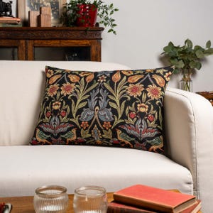 Può includere: Cuscino decorativo rettangolare con sfondo nero e motivo floreale nei toni del rosso, giallo, verde e blu. Il cuscino è posto su un divano bianco. Il design floreale comprende uccelli e fiori stilizzati.