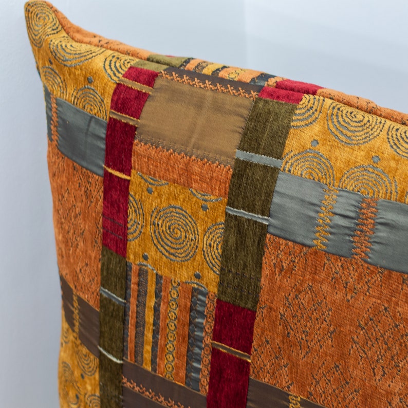 May include: A decorative throw pillow with a patchwork design featuring various shades of brown, orange, red, and green. The fabric has a textured, woven look with geometric patterns and swirls.