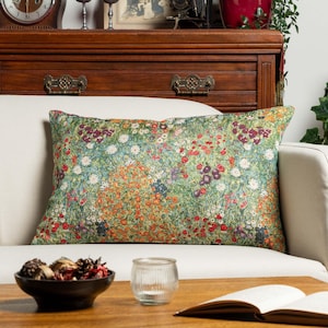 Puede incluir: Un cojín rectangular con un estampado botánico denso y colorido de pequeñas flores en tonos rojo, naranja, blanco y morado sobre un fondo de follaje verde vibrante, colocado sobre un sofá de color claro.