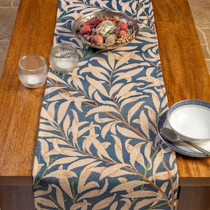 May include: A dining table set with a blue and beige leaf-patterned table runner. A silver bowl filled with decorative items sits on the runner. Two glass votive candle holders and a bowl with a spoon are also present.