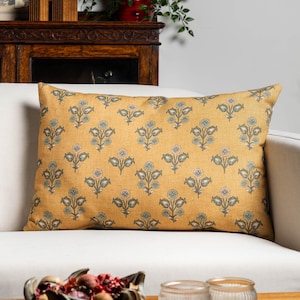 May include: Mustard yellow rectangular cushion featuring a floral pattern in blue, green, and pink. The cushion is placed on a white sofa. The floral design is repeated across the cushion's surface.