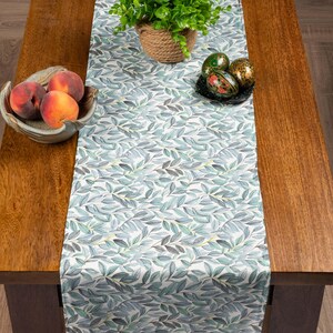 May include: A table runner featuring a repeating pattern of blue-green leaves on a white backdrop. The runner is on a wooden table, with a bowl of peaches and decorative eggs. The table is made of wood.