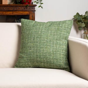 May include: A square, green cushion with a woven texture. The cushion is placed on a white sofa. The cushion's colour is a mix of light and dark green, creating a textured appearance. The cushion is a home decor item.