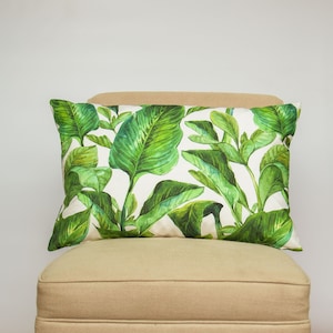 Op de afbeelding: Een wit kussen met een groen tropisch bladmotief. Het kussen ligt op een lichtbruine stoel.