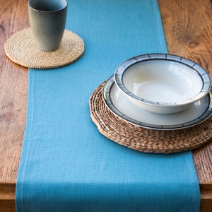 Op de afbeelding: Een blauwe linnen tafelrunner met een geweven placemat en een set van twee keramische borden. De borden zijn wit met een donkergrijze rand.
