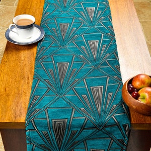 Op de afbeelding: Een turquoise tafelloper met een geometrisch art deco-ontwerp in zilver en zwart. Een kop en schotel met een blauwe rand staan op een houten tafel, samen met een houten kom met fruit. De loper voegt een decoratief tintje toe.