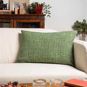 Peut inclure: Un coussin rectangulaire vert texturé repose sur un canapé blanc. Le coussin présente des nuances de vert et un motif subtil. Le canapé est dans une pièce bien éclairée avec une armoire en bois et des objets décoratifs en arrière-plan.