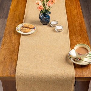 May include: A beige herringbone table runner on a wooden table. A blue vase with pink flowers, two tealight candles, a plate of biscuits, and a teacup and saucer are on the table. The table is made of wood.