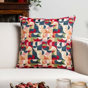 May include: Square cushion with a geometric pattern in red, blue, green, orange, and pink. The cushion is on a white sofa. The design features a repeating pattern of shapes, creating a modern and colourful aesthetic.
