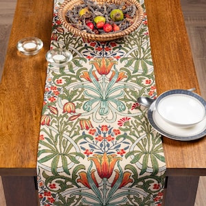 May include: A decorative table runner with a floral pattern in shades of green, orange, and blue. The runner is placed on a wooden table, with a woven basket of fruit and candles visible.