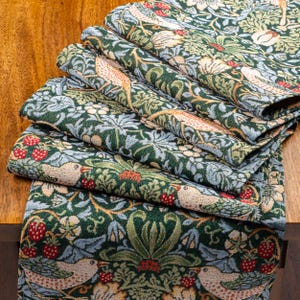 Puede incluir: Un camino de mesa de tela tejida con un patrón repetitivo de pájaros, fresas y follaje en tonos verde oscuro, azul claro, crema y rojo. El textil muestra un diseño botánico denso e intrincado con múltiples pliegues.