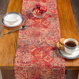 May include: A red and orange patterned table runner with a vintage design. The table is set with a bowl of fruit, a cup of coffee, cookies, a spoon, and a bowl and plate.