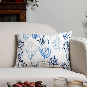 Peut inclure: Coussin rectangulaire décoratif avec un fond blanc et un motif d'algues bleues et grises. Le coussin est posé sur un canapé crème. Le motif d'algues comprend diverses formes et nuances de bleu et de gris.