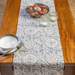 May include: A blue and white patterned table runner with a floral design is laid on a wooden table. A silver bowl filled with decorative items, two glass candle holders, and a bowl with a spoon are on the table.