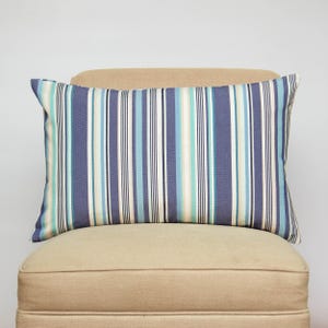 Op de afbeelding: Een rechthoekig kussen met een gestreept patroon in blauwe, turquoise en witte tinten. Het kussen ligt op een beige stoel, wat een kust- of nautisch thema suggereert. De strepen zijn verticaal en gelijkmatig verdeeld.