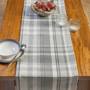 Op de afbeelding: Een grijs en wit geruite tafelloper ligt op een houten tafel. Een decoratieve schaal gevuld met gedroogde potpourri en twee kleine glazen kandelaars staan op de loper. Een kom en lepel staan links.