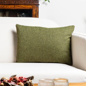 Puede incluir: Un cojín rectangular verde oliva con un patrón de espiga texturizado. El cojín está colocado sobre un sofá blanco. Es un artículo decorativo para la decoración del hogar.