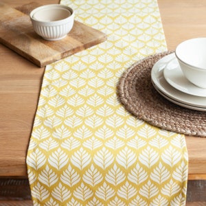 May include: A yellow table runner with a white leaf pattern. The runner is on a wooden table with a brown woven placemat and white dishes.