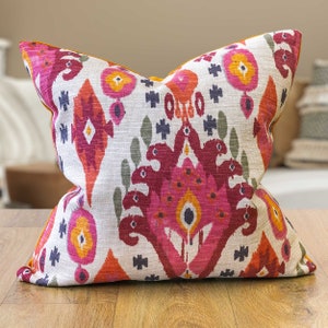 May include: A decorative throw pillow with a white background and a colorful ikat pattern in shades of pink, orange, and blue. The pillow has a mustard yellow border.