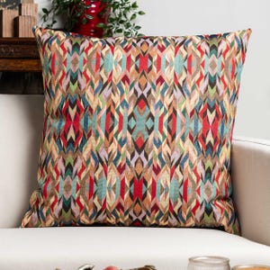 Pode incluir: Almofada decorativa quadrada com um padrão geométrico em tons de vermelho, azul, verde e castanho. A almofada está colocada num sofá branco, mostrando as suas cores vibrantes e o seu design intrincado. O padrão é simétrico.