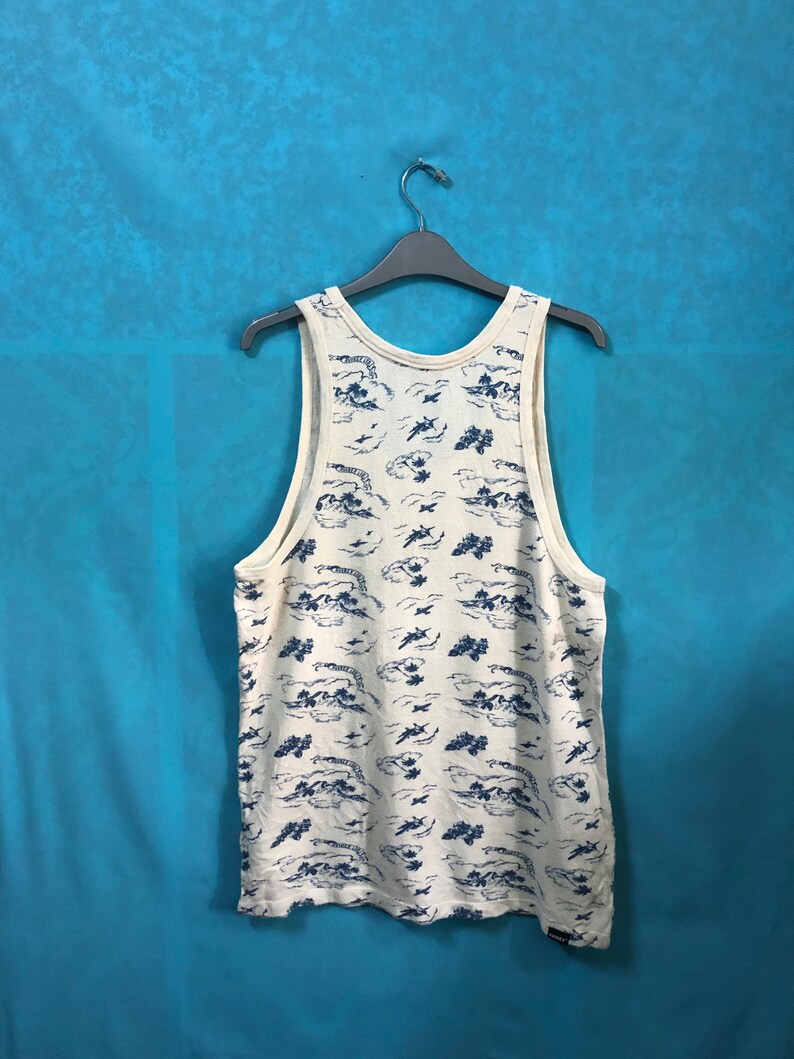 May include: A white sleeveless shirt with a blue and white print of clouds and airplanes. The shirt is hanging on a black hanger against a blue background.