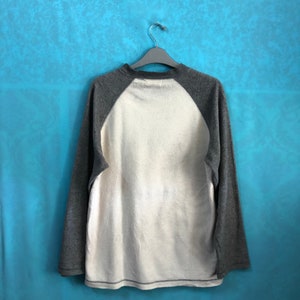 K&ouml;nnte beinhalten: Ein lang&auml;rmliges Shirt mit wei&szlig;em Oberk&ouml;rper und grauen &Auml;rmeln. Die &Auml;rmel sind im Baseball-Stil mit einer kontrastierenden Farbe gehalten. Das Shirt h&auml;ngt an einem Kleiderb&uuml;gel vor einem blauen Hintergrund.