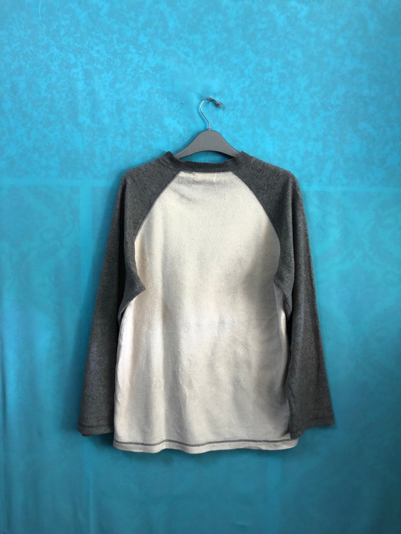 K&ouml;nnte beinhalten: Ein lang&auml;rmliges Shirt mit wei&szlig;em Oberk&ouml;rper und grauen &Auml;rmeln. Die &Auml;rmel sind im Baseball-Stil mit einer kontrastierenden Farbe gehalten. Das Shirt h&auml;ngt an einem Kleiderb&uuml;gel vor einem blauen Hintergrund.