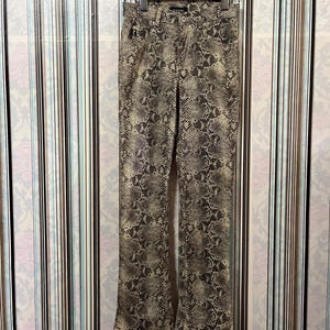 Op de afbeelding: Broek met slangenprint die aan een houten hanger hangt. De broek heeft een uitlopende pijp en een neutraal kleurenpalet van beige, bruin en zwart. De broek is gemaakt van een textuurstof.