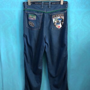 Könnte beinhalten: Dunkelblaue Jeans aus Denim mit grünem Bund und mehreren Patches auf den Gesäßtaschen. Die Patches beinhalten einen "Bull's Club"-Patch, einen "H.A.M.I.E.S"-Patch mit einem Oldtimer und einen runden Patch mit einem Zahnrad-Design.