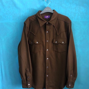 Puede incluir: Una camisa vaquera de manga larga de color marrón con cuello de punta, cierre de botones y dos bolsillos en el pecho con tachuelas en forma de diamante. La camisa tiene una etiqueta morada con el texto "REPRESENT"