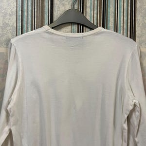 May include: A white long-sleeve shirt hanging on a gray hanger. The shirt has a crew neck and is made of a soft, plain fabric. The background features a striped wallpaper.
