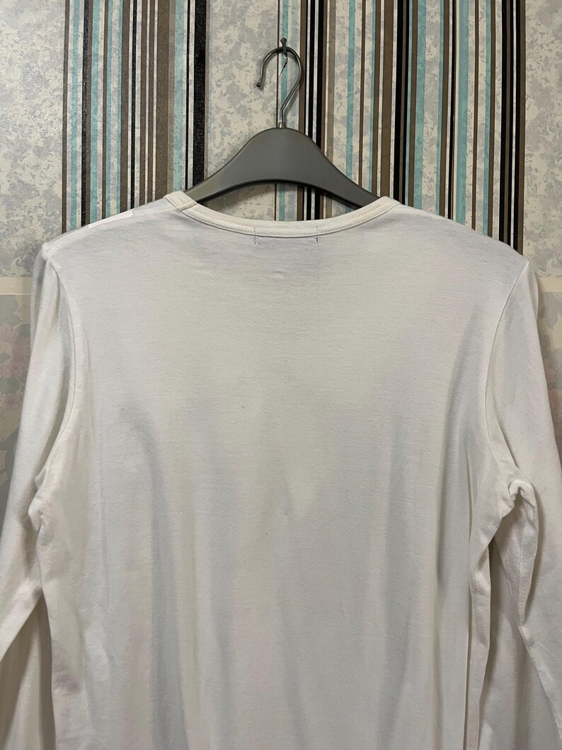 May include: A white long-sleeve shirt hanging on a gray hanger. The shirt has a crew neck and is made of a soft, plain fabric. The background features a striped wallpaper.