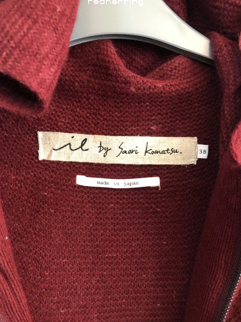 Puede incluir: Un c&aacute;rdigan de punto color burdeos con cierre de cremallera. La etiqueta dice "il by Saori Komatsu." y "made in japan."