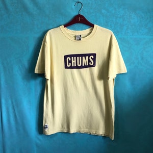 Peut inclure: Un t-shirt jaune clair avec le mot "CHUMS" dans un rectangle violet foncé sur le devant. Le t-shirt est suspendu à un cintre en bois sur un fond bleu. Un petit logo est visible sur la manche supérieure gauche.