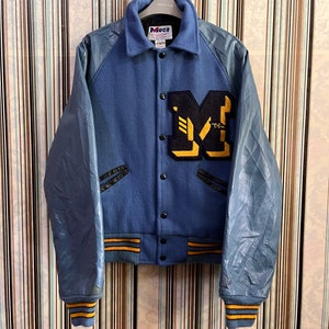 Könnte beinhalten: Eine blaue und schwarze Varsity-Jacke mit Lederärmeln. Die Jacke hat einen großen gelben und schwarzen Buchstaben "M" auf der Vorderseite. Die Jacke hat einen gelben und schwarzen Streifenbesatz an den Ärmelbündchen und am Saum.