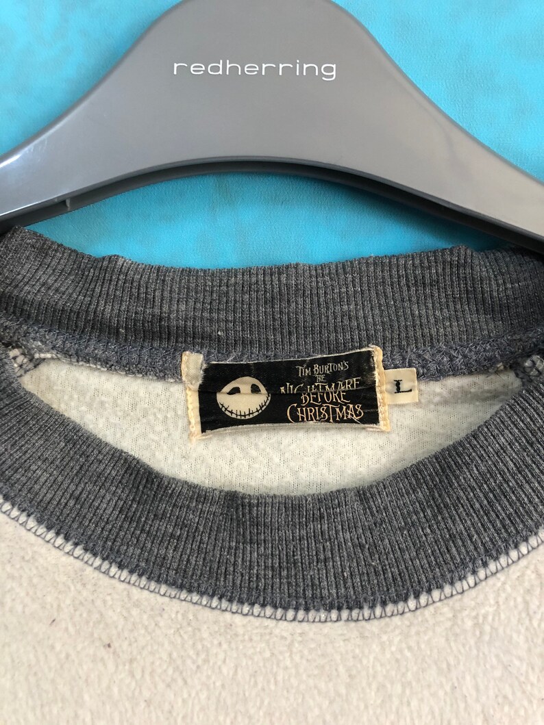 K&ouml;nnte beinhalten: Ein wei&szlig;-graues Sweatshirt mit einem schwarz-wei&szlig;en Etikett, auf dem "Tim Burton's The Nightmare Before Christmas" steht. Das Etikett zeigt eine schwarz-wei&szlig;e Illustration von Jack Skellingtons Gesicht.