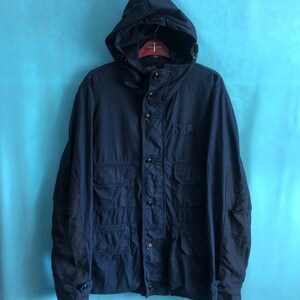 May include: Navy blue hooded jacket with multiple front pockets and button closures. The jacket features a drawstring hood and a classic, utilitarian design. The jacket is hanging on a wooden hanger against a light blue background.